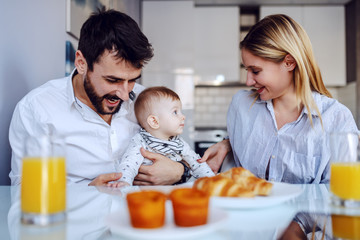 Young happy parents sitting in dining room with their adorable baby boy. Man holding baby. On table is breakfast.