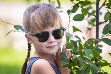 Young pretty child girl in fashionable sun glasses hugging green small tree in summer outdoors.