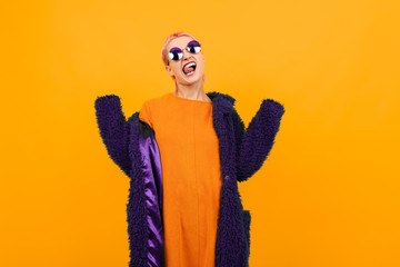 Extraordinary beautiful woman with short pink hair in dark coat and sunglasses smiles isolated on orange background