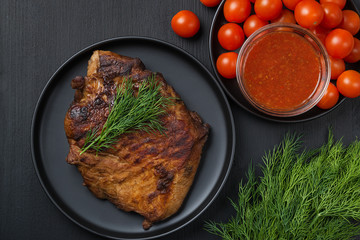 Grilled pork steak, tomatoes and sauce in a plate on a black table. View from above.