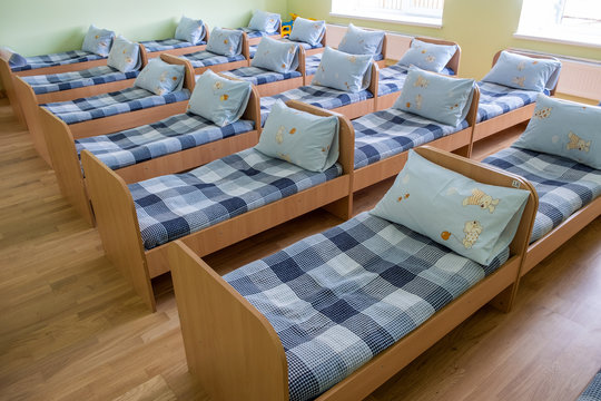 Many Small Beds With Fresh Linen In Daycare Preschool Empty Bedroom Interior For Comfortable Afternoon Nap Of The Kids.