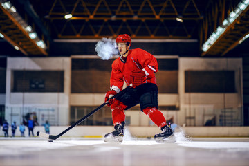 Dedicated strong hockey player in red uniform with protective helmet on head skating and going to...