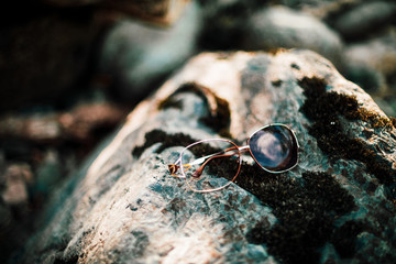 Broken sunglasses on stone with moss in sunlight. Lost thing on mossy boulder in sunny day. Glare on shiny thing. Nonsense happened. Lost sunglasses on rock close-up. Things break, life goes on.