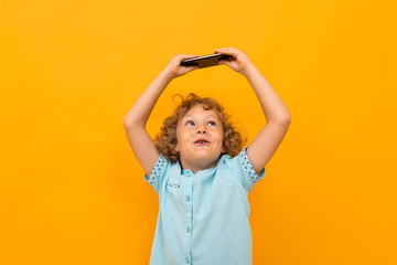 Little boy with curly hair in blue shirt and shorts play phone games isolated on yellow background