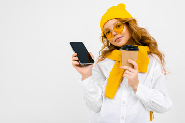 Closeup portrait of a cute caucasian teenager girl in a winter hat and scarf with a phone with a breadboard in his hand and a paper cup on a white background