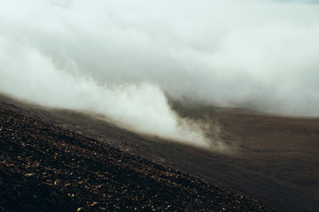 View from stony mountainside to highland valley within clouds. Atmospheric darker minimalist alpine landscape with low clouds in mountain valley. Wonderful mountain scenery with thick clouds on ground