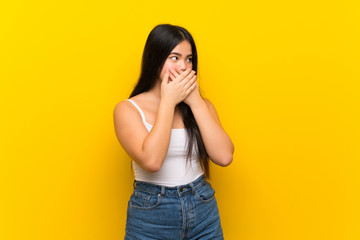 Young teenager Asian girl over isolated yellow background covering mouth and looking to the side