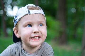 Cute little caucasian baby boy with blond hair and blue eyes smiling. Outdoor, green park or forest around there. Casual wear, cap backwards.