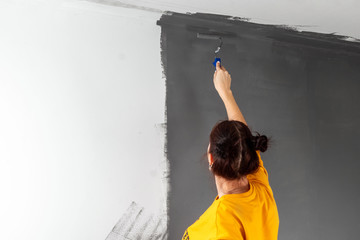 Obraz premium beautiful girl in a yellow sweater paints a wall in gray paint. Room painting, repair, design. Copy space.