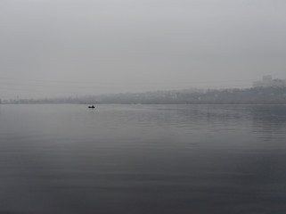 Fishermen on an inflatable boat in the middle of the river against the background of the city's high-rise buildings. Foggy weather, weekend, hobby.