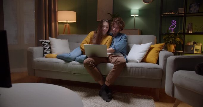 Young Caucasian happy in love couple resting on the couch and hugging while watching something on laptop computer together late at night and smiling. Indoors.