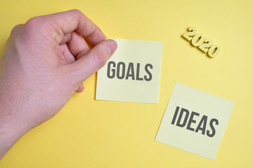The hand sticks a sticker with the inscription of goals and ideas next to wooden figures 2020 on a yellow background. Top view.