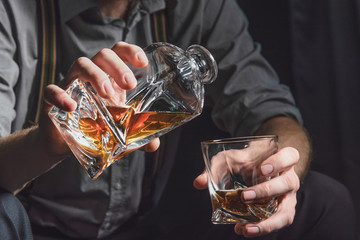 Close-up of the hands of a young man who pours alcohol from a decanter into a glass, on a dark...