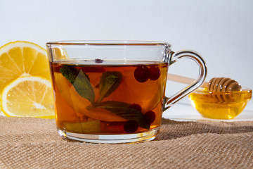 Glass cup with tea, lemon berries and mint on a mat, against the background of lemon and honey in a glass bowl.