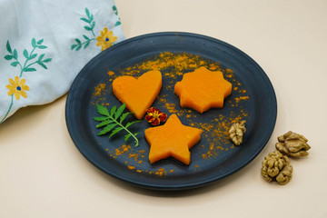 Baked pumpkin on a plate of black ceramics on pastel background. Pumpkin portions in the form of heart, star and flower. The dish is decorated with walnuts and marigold. An embroidered towel nearby.