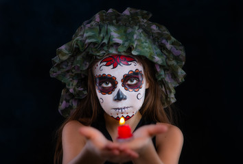 Little beautiful girl with a sugar skull makeup holds a candle in her hands on a dark background. ...