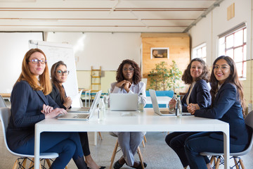 Businesswomen sitting at table and smiling at camera. Group of professional cheerful multiethnic businesswomen sitting around table in meeting room. Women in business concept