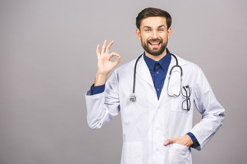 Portrait of cheerful smiling young doctor with stethoscope over neck in medical coat standing against isolated gray background. Ok sign.