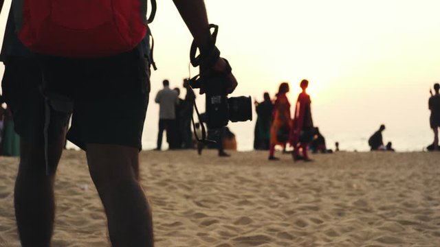 Medium closeup of a man wearing shots walking with a DSLR camera on a beach during sunset