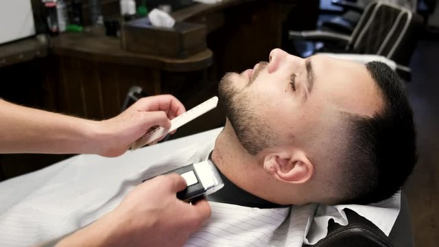 A client has a beard. He has dark hair. A barber is working in salon.