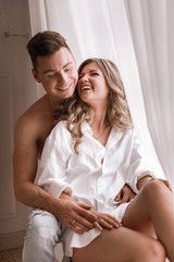 loving young couple enjoying morning at home near the window on Valentine's Day. girl in white shirt and guy half naked having fun together