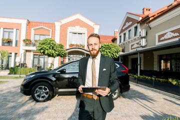 Young businessman or sales manager in dark suit standing outdoor on the yard of business center with new black car on background