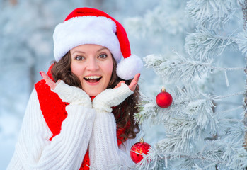 Beautiful girl in the winter forest in a Santa hat near a dressed-up Christmas tree. In white and red colors