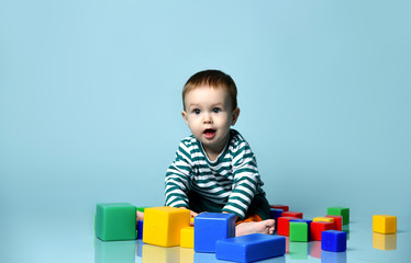 Obraz premium Little baby boy in stylish casual clothing barefoot sitting on floor and playing with colorful toy cubes over blue wall background