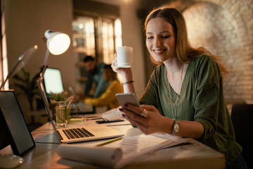 Smiling freelancer texting on cell phone and drinking coffee in the office.
