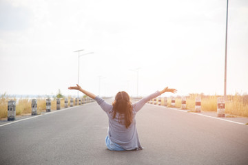 The back view of a young woman sitting on the road, looking far away and hands up.