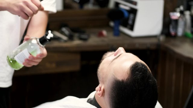 A barber holds a spray. A client closed his eyes. The men are in the barbershop.