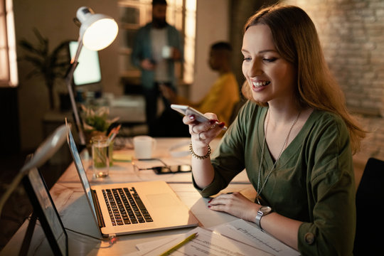 Young Smiling Freelancer Using Smart Phone While Working Late At The Office.