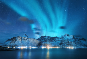 Fototapeta na wymiar Aurora borealis over the sea, snowy mountains and city lights at night. Northern lights in Lofoten islands, Norway. Blue starry sky with polar lights. Winter landscape with aurora reflected in water