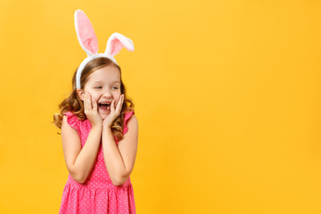 Funny little girl in Easter bunny ears on a yellow background. The child opened his mouth in surprise and looks at the empty space