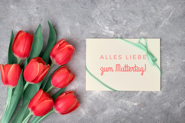 "Alles Liebe zum Muttertag", that means in German "All the love on Mother's day". Text on paper card. Flat lay with red tulips, green decorative flowers and paper card on grey textured background.