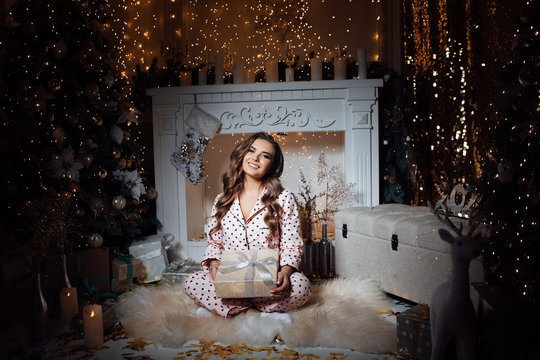 Lovely girl in a photo studio. Girl with Christmas gift. Beautiful woman in in pink pajamas in polka dots in a beautiful bright room with decoration near Christmas tree and fireplace with garlands lan