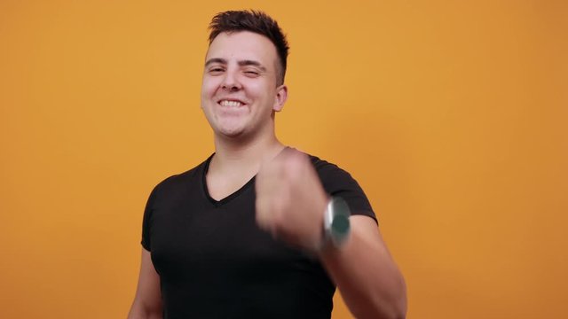 Cheerful young man wearing black shirt isolated on orange background in studio waving his fist, dancing, smiling. People sincere emotions, lifestyle concept.