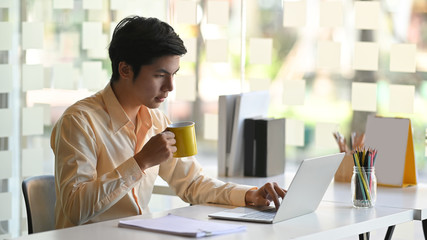 Businessman holding a coffee cup and looking at laptop when sitting at the work desk in the modern...