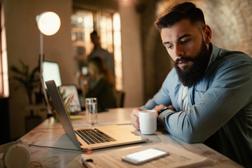 Fototapeta premium Young businessman reading text message on mobile phone while working late in the office.
