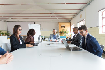 Group of cheerful managers during morning briefing. Smiling workers sitting at table and working with laptops. Business meeting concept