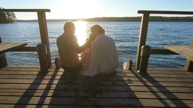 Couple with child sitting on a wooden jetty at sunset near the lake at sunset after the sauna in summer white night in Finland.