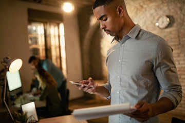 Black businessman text messaging on cell phone while working late in the office.