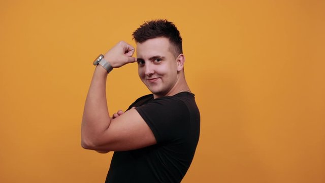 Handsome caucasian young man wearing black shirt isolated on orange background in studio looking at camera, showing muscules, having nice clock. People sincere emotions, lifestyle concept.