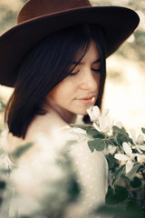 Sensual portrait of beautiful hipster woman in hat smelling  white flowers in spring. Stylish calm boho girl posing in blooming tree with flowers in sunny spring park. Copy space