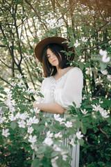 Stylish boho woman in hat posing in blooming tree with white flowers in sunny spring park. Calm portrait of beautiful hipster girl standing in white blooms in spring