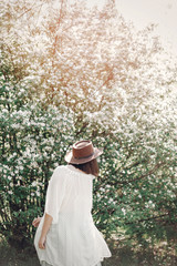 Happy boho woman in hat having fun in sunny light near white blooming tree in spring park. Stylish hipster girl enjoying spring and dancing. Atmospheric moment of happiness