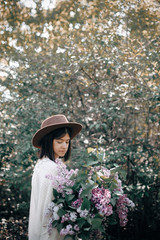 Fototapeta na wymiar Stylish boho woman in hat holding lilac flowers bouquet in sunny spring park. Calm portrait of beautiful hipster girl standing with purple lilac in spring garden. Copy space