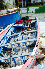 Fototapeta na wymiar Old and broken little small fishing boat. Rotting wood but still preserved color lines. White blue and red. The boat is parked in an empty pool. served as a decoration in front of the restaurant.