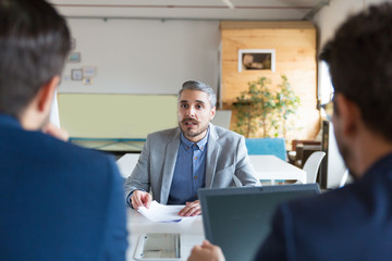 Confident project manager talking to subordinates. Back view of workers sitting at table and listening boss. Business meeting concept