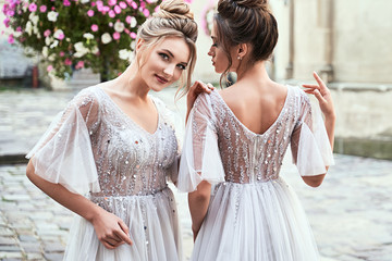 Two beautiful bridesmaids ladies in gorgeous elegant stylish light grey silver floor length open back dresses in old beautiful European city on a wedding day. - obrazy, fototapety, plakaty